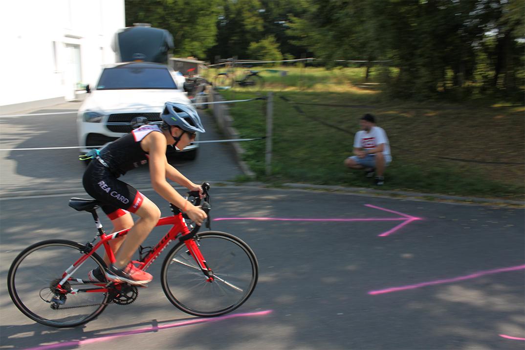 Junge fährt auf einem roten Rennrad, während ein Mann im Hintergrund auf dem Boden sitzt. Pinke Markierungen auf dem Asphalt.