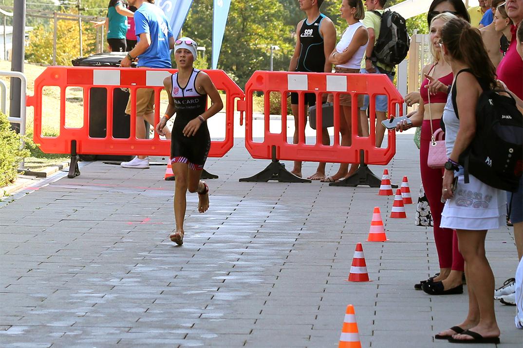 Junge Athletin in Wettkampf-Ausrüstung läuft an Zuschauern und Barrieren vorbei, orange Pylonen markieren die Strecke.
