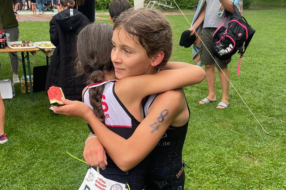 Zwei Mädchen umarmen sich, eines hält ein Stück Wassermelone in der Hand, im Hintergrund eine Veranstaltung im Freien.