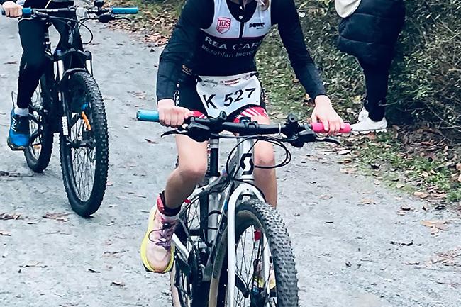 Zwei Kinder fahren auf Mountainbikes einen Waldweg entlang, Zuschauer stehen am Rand und beobachten das Rennen.