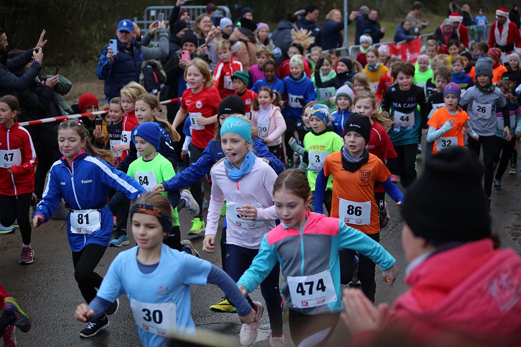 Gruppierung junger Läuferinnen und Läufer beim Start eines Kinderlaufs mit verschiedenen Sporttrikots und Aufregung im Publikum.