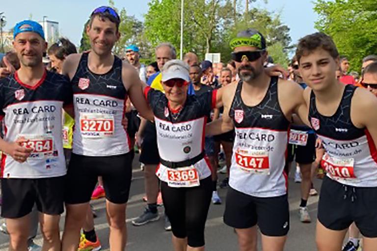 Gruppe von fünf Läufern in Wettkampfkleidung vor dem Start einer Laufveranstaltung mit Zuschauer im Hintergrund.