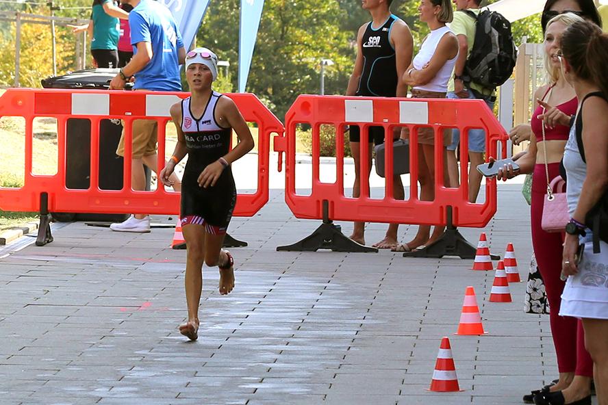Junge Athletin in Wettkampf-Ausrüstung läuft an Zuschauern und Barrieren vorbei, orange Pylonen markieren die Strecke.