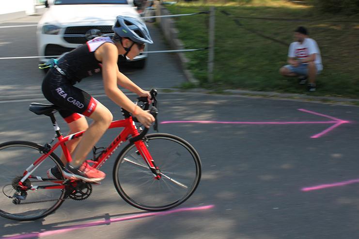 Junge f&auml;hrt auf einem roten Rennrad, w&auml;hrend ein Mann im Hintergrund auf dem Boden sitzt. Pinke Markierungen auf dem Asphalt.
