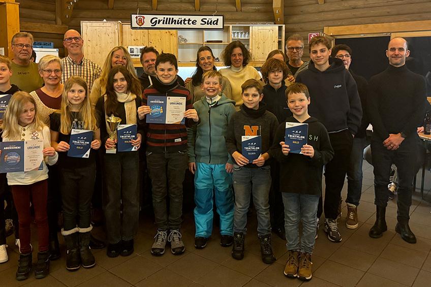 Gruppenbild von Teilnehmern einer Triathlon-Veranstaltung mit Urkunden und Pokalen in festlich dekoriertem Raum.