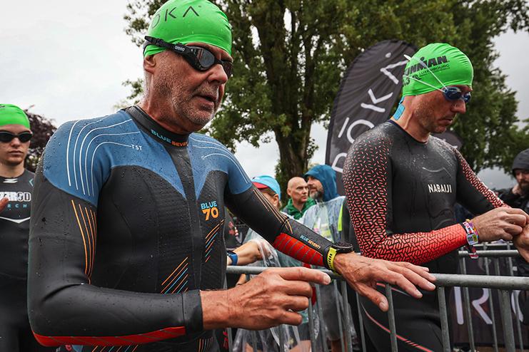 Zwei Triathleten in Neoprenanz&uuml;gen warten am Start, umgeben von Zuschauern und Fahnenschwenkern bei regnerischem Wetter.