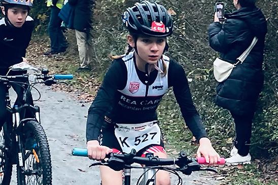 Zwei Kinder fahren auf Mountainbikes einen Waldweg entlang, Zuschauer stehen am Rand und beobachten das Rennen.
