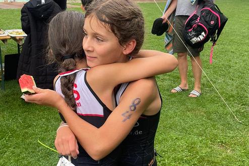 Zwei Mädchen umarmen sich, eines hält ein Stück Wassermelone in der Hand, im Hintergrund eine Veranstaltung im Freien.