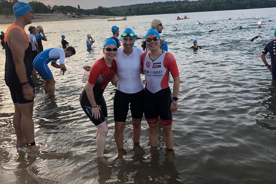 Drei Athletinnen im Wasser lächeln gemeinsam, während im Hintergrund weitere Schwimmer und eine weite Landschaft zu sehen sind.
