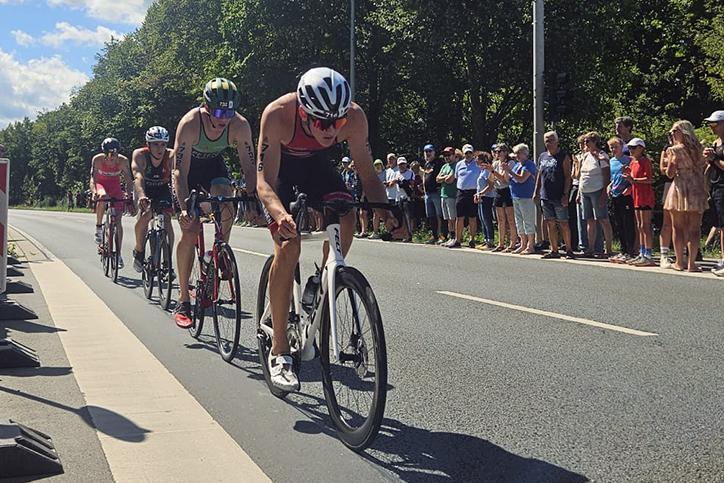 Sechse Triathleten radeln in einem Wettbewerb auf einer Stra&szlig;e, umgeben von Zuschauern unter blauem Himmel.