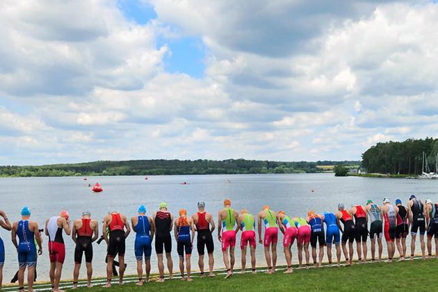 Gruppierung von Triathleten am Seeufer, bereit zum Start in den Wettkampf, mit Zuschauern im Hintergrund.