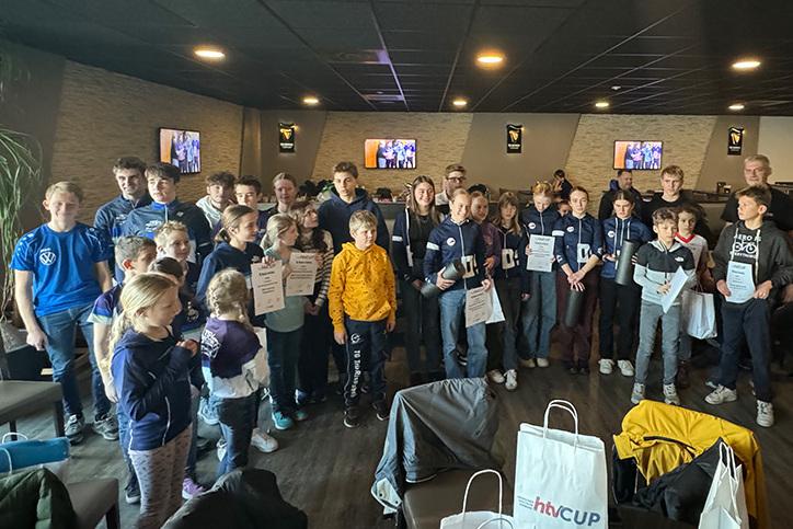 Gruppe von Kindern und Jugendlichen h&auml;lt Zertifikate in einem Veranstaltungsraum mit Fernsehern im Hintergrund.