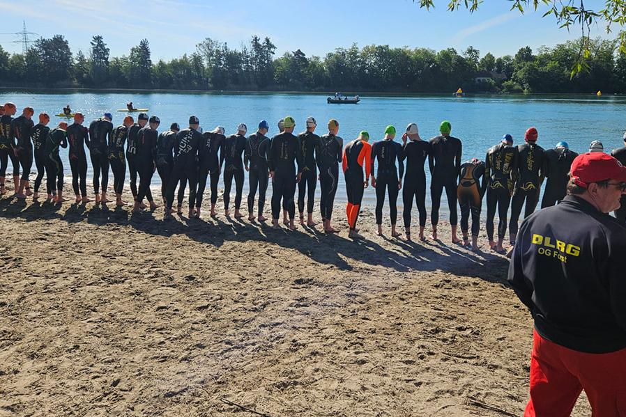 Gruppierung von Schwimmern in Neoprenanzügen am Ufer eines Gewässers, im Hintergrund Boote und eine sonnige Landschaft.