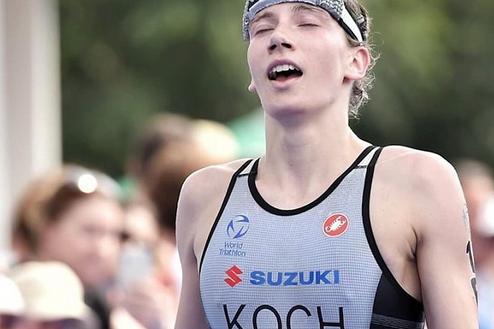 Triathlet überquert die Ziellinie mit blauem Zielband, sichtbar erschöpft, im Hintergrund jubelnde Zuschauer.