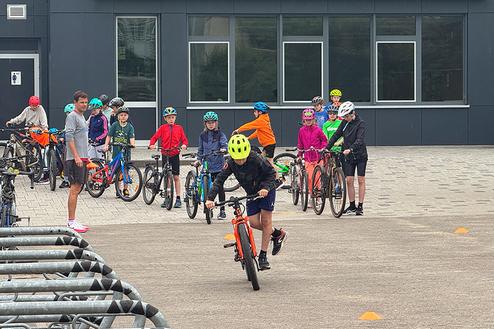 Gruppe von Kindern in bunten Helmen mit Fahrrädern auf einem Platz vor einem Gebäude, während ein Junge fährt.