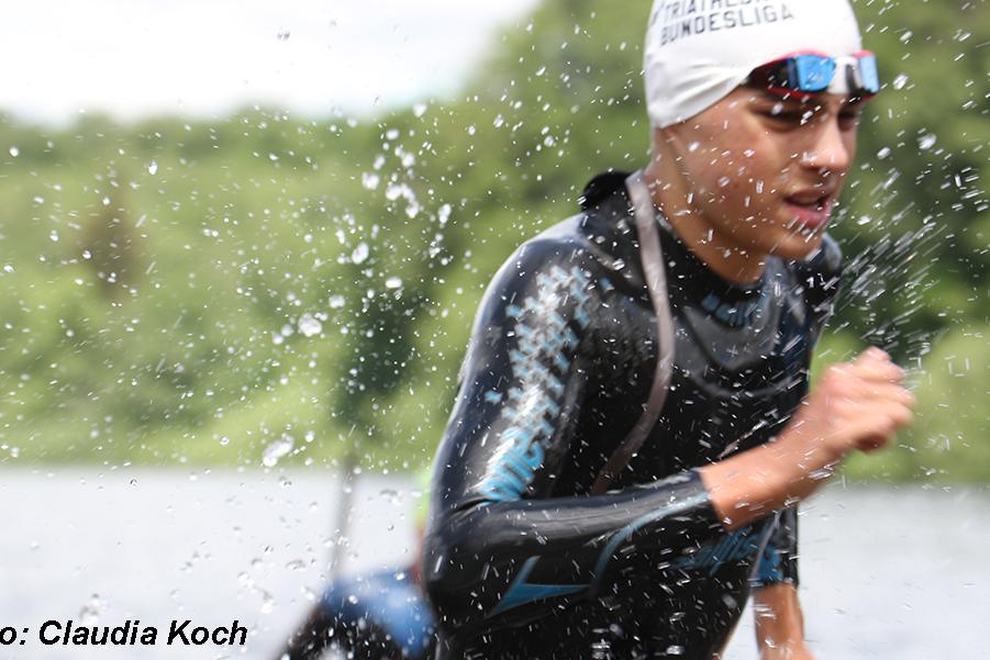 Junger Triathlet läuft nach dem Schwimmen aus einem Gewässer, Wasser spritzt um ihn herum.