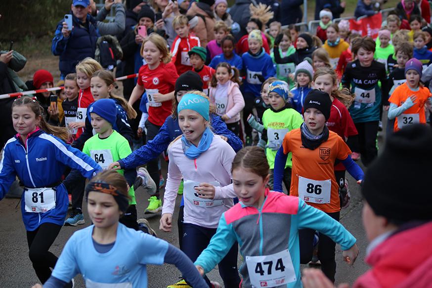 Gruppierung junger Läuferinnen und Läufer beim Start eines Kinderlaufs mit verschiedenen Sporttrikots und Aufregung im Publikum.