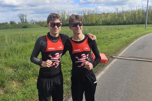 Zwei junge Männer in Sportkleidung stehen lächelnd auf einer Wiese neben einer Landstraße und posieren für ein Foto.