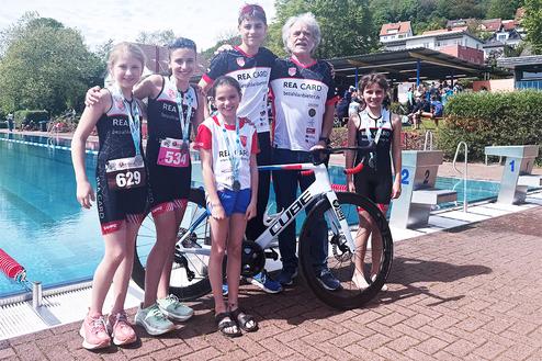 Sechs Kinder in Sportbekleidung stehen neben einem Rad vor einem Schwimmbecken bei einem Triathlon-Event.
