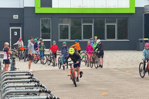 Gruppe von Kindern in bunten Helmen mit Fahrrädern auf einem Platz vor einem Gebäude, während ein Junge fährt.