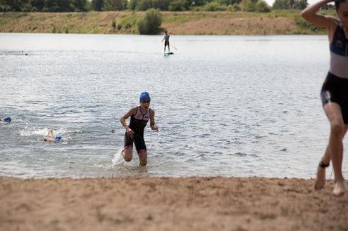 Ein Triathlet in Neoprenanzug läuft aus dem Wasser, während Schwimmer im Hintergrund weiter trainieren.
