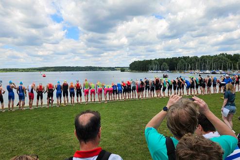 Gruppierung von Triathleten am Seeufer, bereit zum Start in den Wettkampf, mit Zuschauern im Hintergrund.