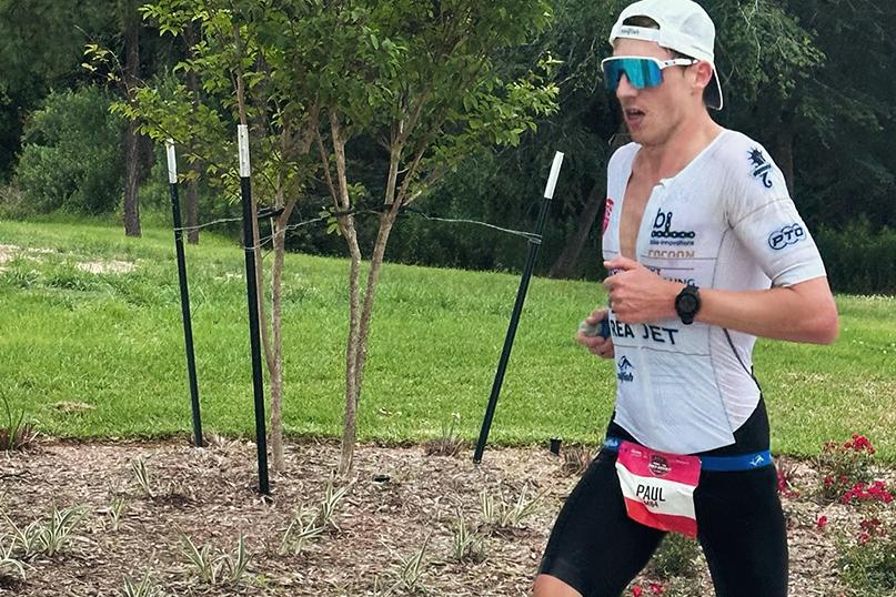 Läufer in sportlicher Kleidung mit Sonnenbrille rennt auf einem gepflasterten Weg, umgeben von Bäumen und Sträuchern.