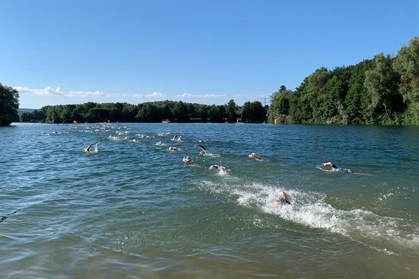 Gruppenschwimmer im klaren Wasser eines Sees, umgeben von Bäumen und blauem Himmel, einige mit Schwimmbojen.