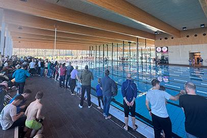 Blick auf ein Schwimmbecken mit Zuschauern und Schwimmern, umgeben von modernen Holzelementen und gro&szlig;en Fenstern.