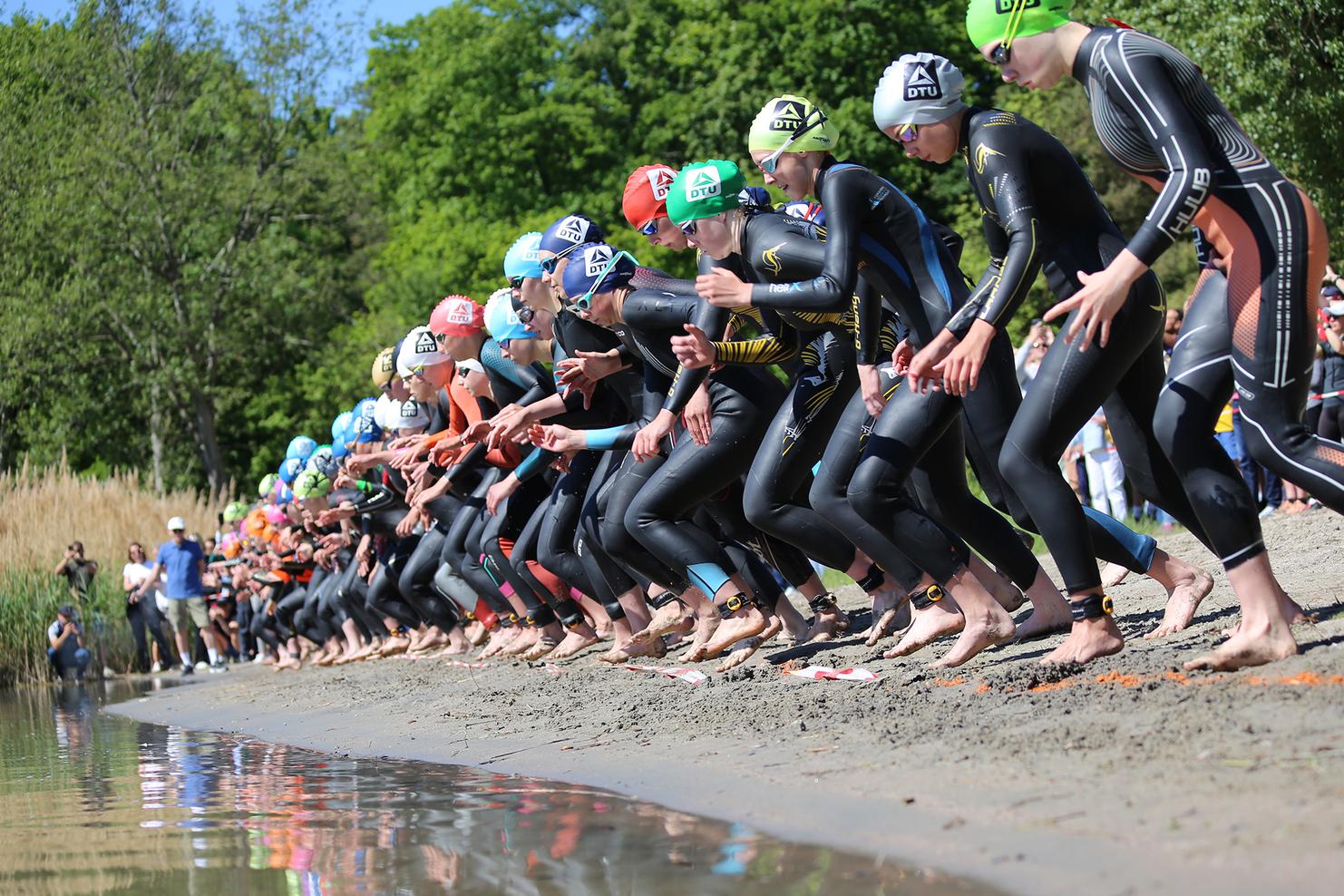 Sportler in Neoprenanzügen stehen am Ufer eines Gewässers, bereit zum Start eines Wettkampfs. Zuschauer beobachten auf der Seite.