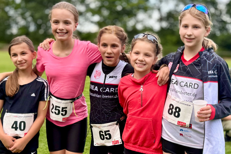 F&uuml;nf M&auml;dchen in Sportoutfits stehen auf einer Wiese, l&auml;cheln und posieren nach einem Wettkampf.