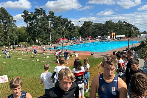Gruppen junger Schwimmer stehen vor einem Wettkampfbecken, umgeben von Zuschauern und Festzelten unter einem blauen Himmel.