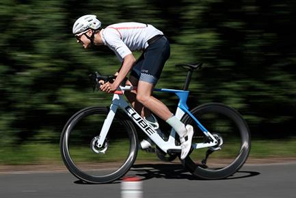 Rennradfahrer in Fahrt, gekleidet in Sportbekleidung, fährt schnell auf einer Straße, umgeben von Bäumen.