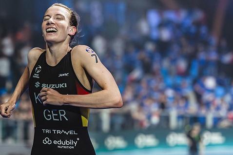 Lächelnde Athletin in Sportkleidung endet einen Triathlon, umgeben von jubelndem Publikum in einer Halle.