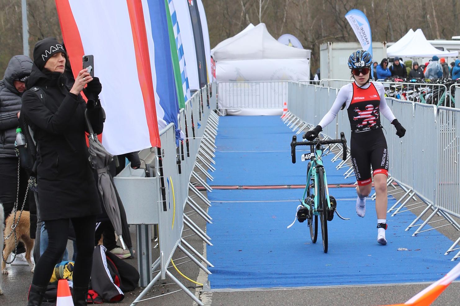 Athlet in Triathlonsportkleidung schiebt ein Rennrad auf einer blauen Fläche, während Zuschauer in der Umgebung stehen.