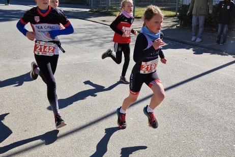 Drei Kinder in Laufschuhen rennen auf einer Straße, alle mit Startnummern auf ihren Sportanzügen.