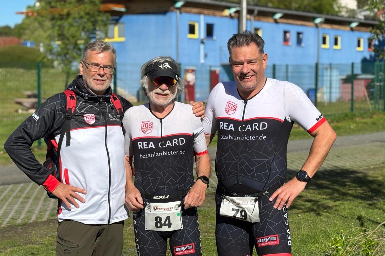 Drei Männer in Sportbekleidung stehen vor einem blauen Gebäude, bereit für einen Wettkampf im Triathlon.