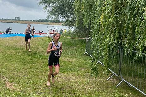 Mädchen in Wettkampfkleidung läuft am Wasser entlang, während im Hintergrund weitere Teilnehmer zu sehen sind.