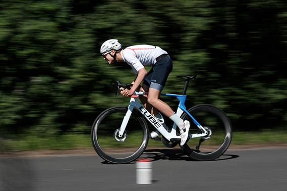 Rennradfahrer in Fahrt, gekleidet in Sportbekleidung, fährt schnell auf einer Straße, umgeben von Bäumen.