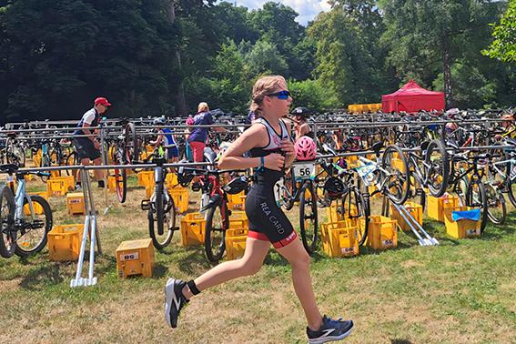 Junge Sportlerin in Wettkleidung läuft durch eine Transitionzone mit Fahrrädern und gelben Transportboxen.