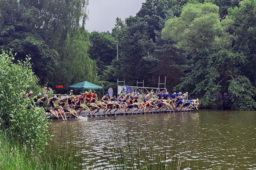 Sportler springen vom Steg ins Wasser bei einem Triathlon-Event in einem bewaldeten Umfeld. Grüne Vegetation umgibt den See.