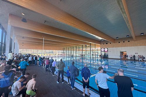 Blick auf ein Schwimmbecken mit Zuschauern und Schwimmern, umgeben von modernen Holzelementen und großen Fenstern.