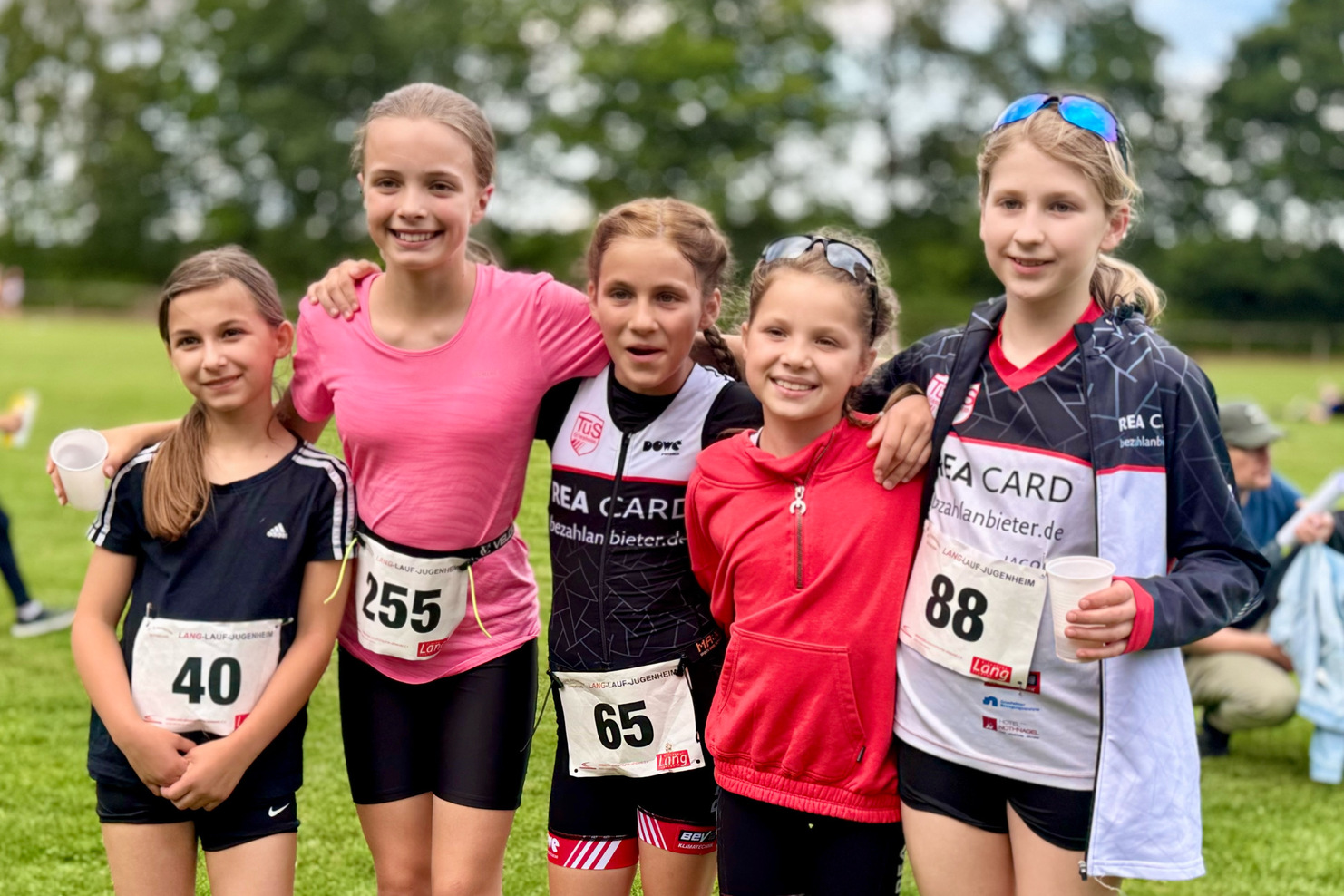 Fünf Mädchen in Sportoutfits stehen auf einer Wiese, lächeln und posieren nach einem Wettkampf.
