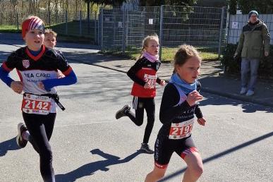 Drei Kinder in Laufschuhen rennen auf einer Straße, alle mit Startnummern auf ihren Sportanzügen.