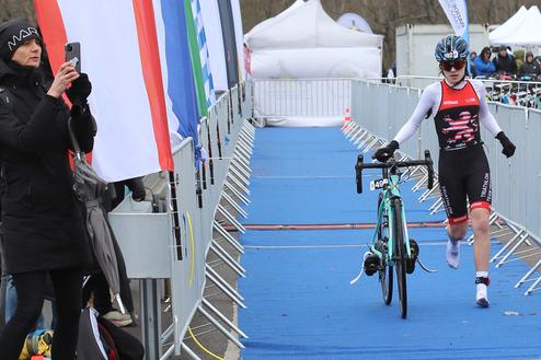 Athlet in Triathlonsportkleidung schiebt ein Rennrad auf einer blauen Fläche, während Zuschauer in der Umgebung stehen.