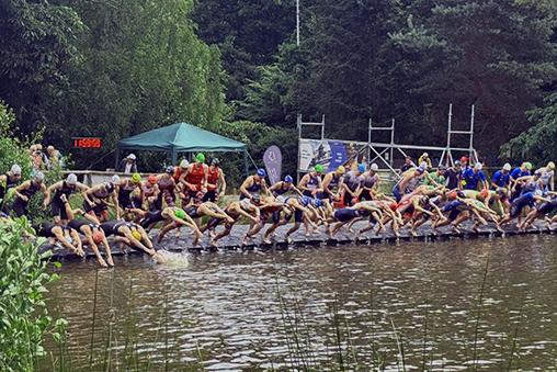 Sportler springen vom Steg ins Wasser bei einem Triathlon-Event in einem bewaldeten Umfeld. Grüne Vegetation umgibt den See.