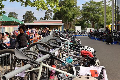 Ansicht eines Fahrradparkplatzes mit mehreren Fahrr&auml;dern, einer Warteschlange von Menschen und B&auml;umen im Hintergrund.