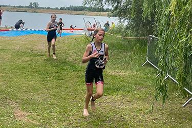 Mädchen in Wettkampfkleidung läuft am Wasser entlang, während im Hintergrund weitere Teilnehmer zu sehen sind.