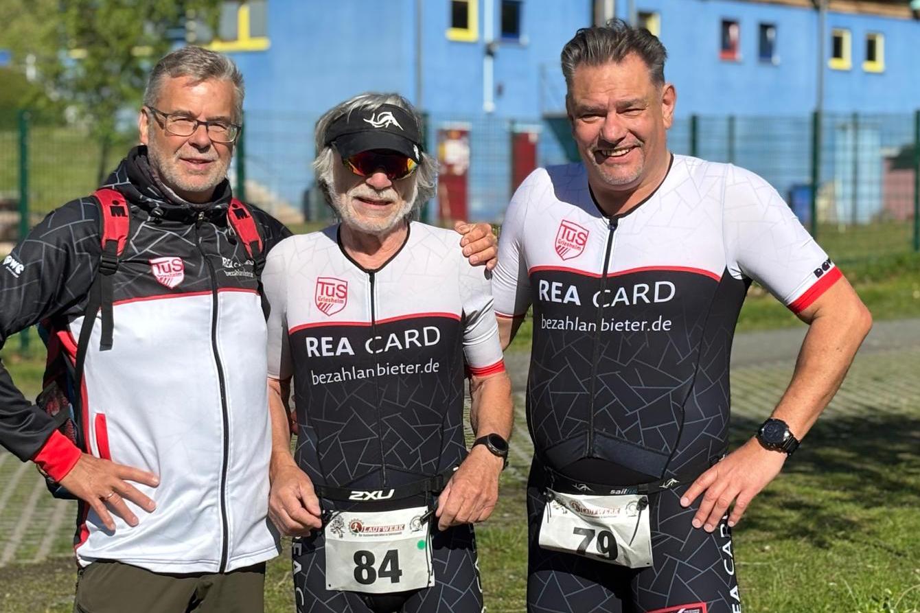 Drei Männer in Sportbekleidung stehen vor einem blauen Gebäude, bereit für einen Wettkampf im Triathlon.