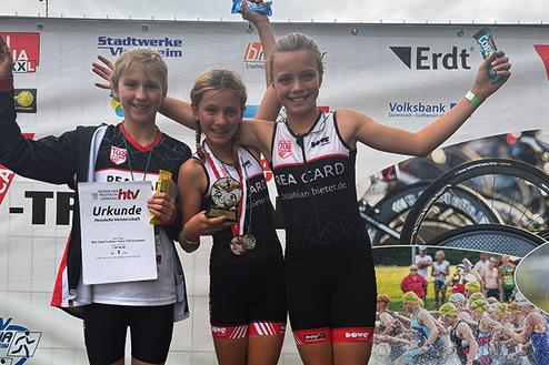 Drei Mädchen in Sportkleidung posieren stolz mit Medaillen und Urkunden vor einem Veranstaltungsbanner.